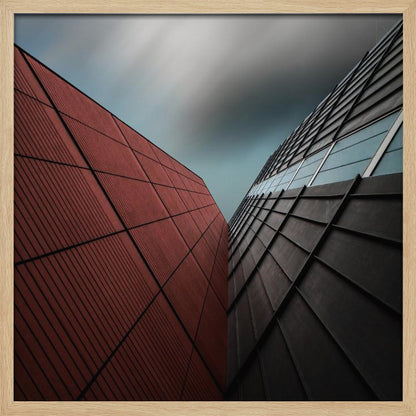A dramatic low-angle photograph of two modern buildings with contrasting facades. On the left, a building is covered in deep red square panels. On the right, a darker building features black angular tiles and glass windows. The sky above is a moody, blurred grey. Print
