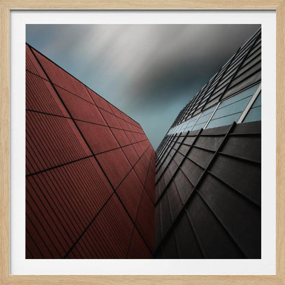 A dramatic low-angle photograph of two modern buildings with contrasting facades. On the left, a building is covered in deep red square panels. On the right, a darker building features black angular tiles and glass windows. The sky above is a moody, blurred grey. Print