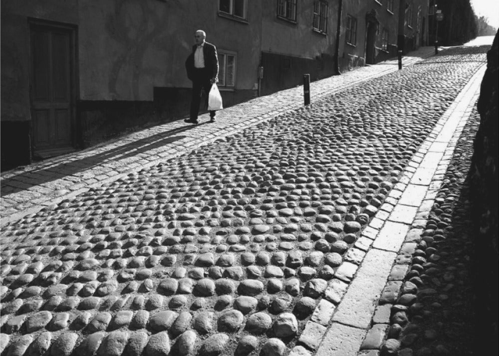 A framed, black and white photograph capturing an elderly man in a suit walking up a steep cobblestone street. The man carries a white bag and casts a long shadow behind him in the harsh sunlight. The street is lined with old, multi-story buildings, creating a classic European city scene. Poster