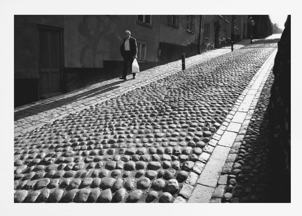 A framed, black and white photograph capturing an elderly man in a suit walking up a steep cobblestone street. The man carries a white bag and casts a long shadow behind him in the harsh sunlight. The street is lined with old, multi-story buildings, creating a classic European city scene. Poster