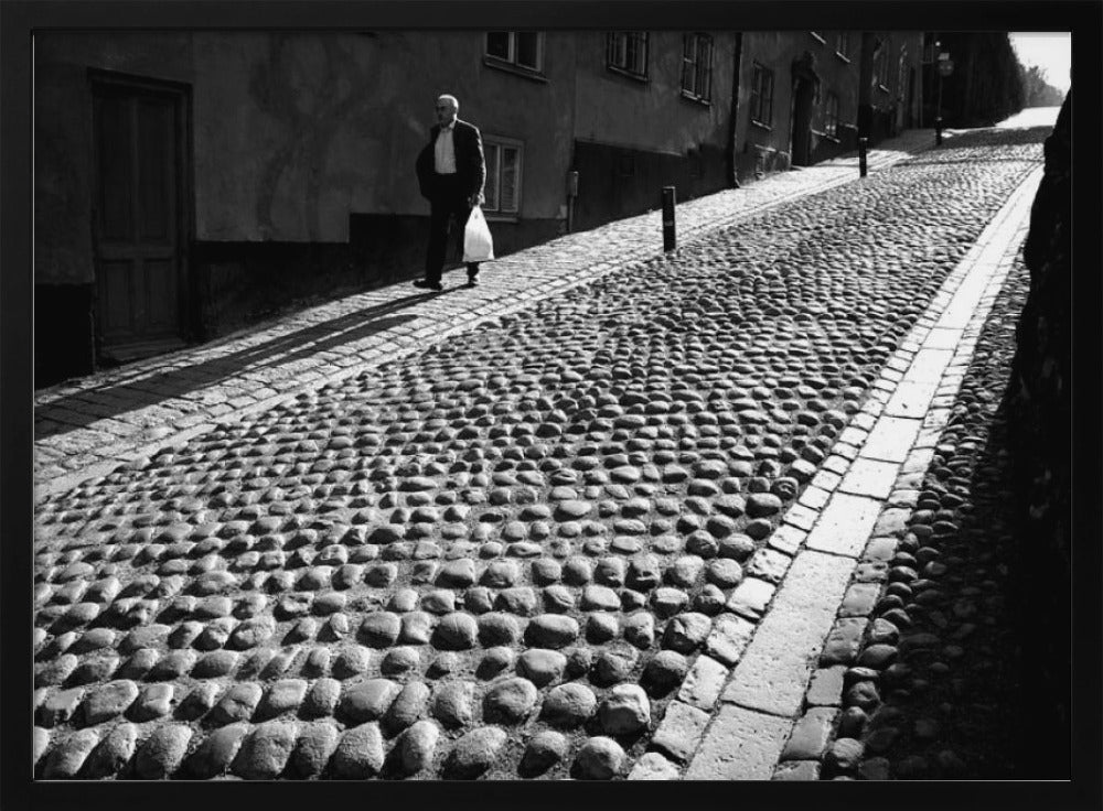 A framed, black and white photograph capturing an elderly man in a suit walking up a steep cobblestone street. The man carries a white bag and casts a long shadow behind him in the harsh sunlight. The street is lined with old, multi-story buildings, creating a classic European city scene. Poster