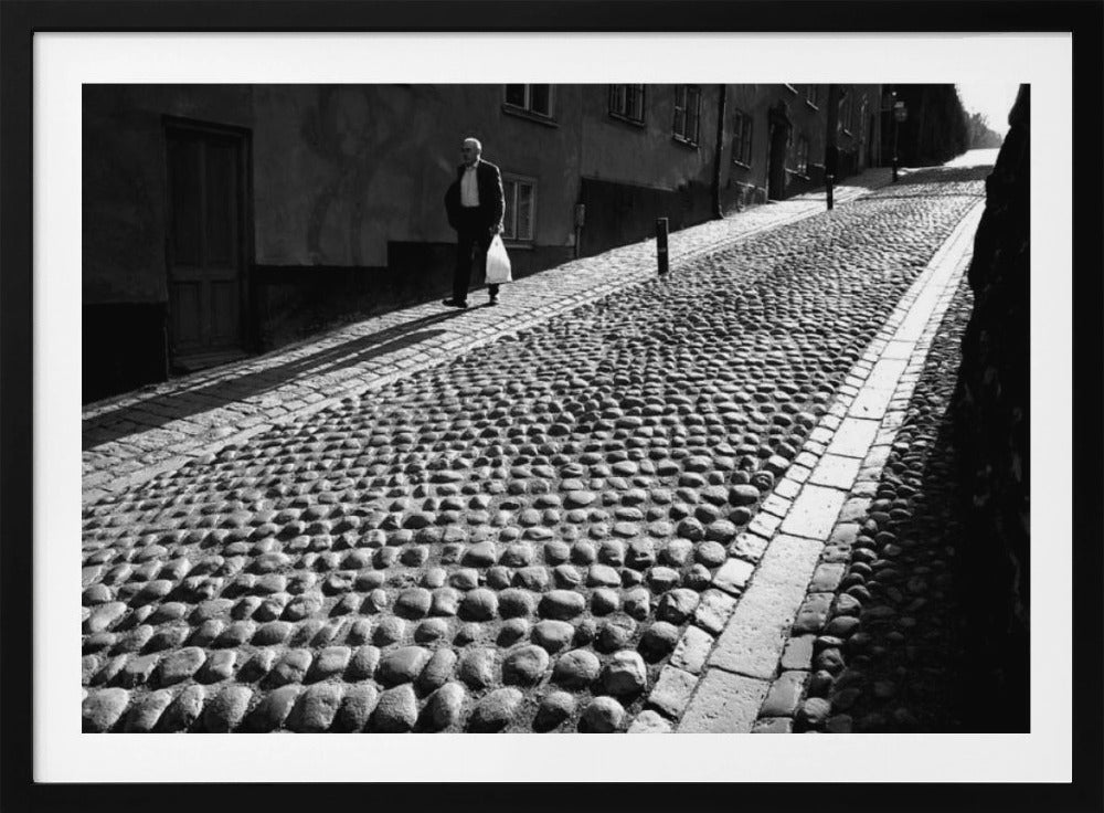 A framed, black and white photograph capturing an elderly man in a suit walking up a steep cobblestone street. The man carries a white bag and casts a long shadow behind him in the harsh sunlight. The street is lined with old, multi-story buildings, creating a classic European city scene. Poster