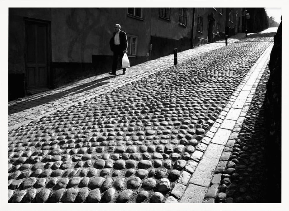A framed, black and white photograph capturing an elderly man in a suit walking up a steep cobblestone street. The man carries a white bag and casts a long shadow behind him in the harsh sunlight. The street is lined with old, multi-story buildings, creating a classic European city scene. Poster