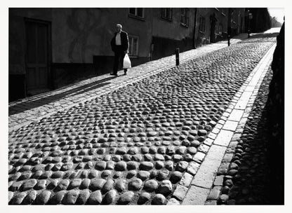 A framed, black and white photograph capturing an elderly man in a suit walking up a steep cobblestone street. The man carries a white bag and casts a long shadow behind him in the harsh sunlight. The street is lined with old, multi-story buildings, creating a classic European city scene. Poster