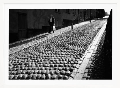 A framed, black and white photograph capturing an elderly man in a suit walking up a steep cobblestone street. The man carries a white bag and casts a long shadow behind him in the harsh sunlight. The street is lined with old, multi-story buildings, creating a classic European city scene. Poster