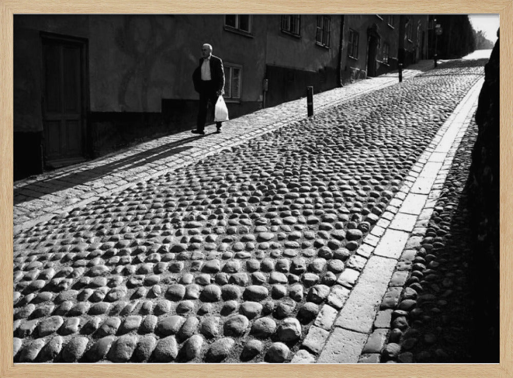 A framed, black and white photograph capturing an elderly man in a suit walking up a steep cobblestone street. The man carries a white bag and casts a long shadow behind him in the harsh sunlight. The street is lined with old, multi-story buildings, creating a classic European city scene. Poster