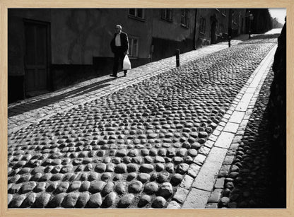 A framed, black and white photograph capturing an elderly man in a suit walking up a steep cobblestone street. The man carries a white bag and casts a long shadow behind him in the harsh sunlight. The street is lined with old, multi-story buildings, creating a classic European city scene. Poster