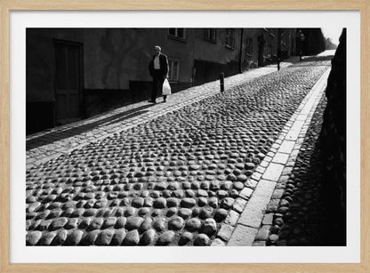 A framed, black and white photograph capturing an elderly man in a suit walking up a steep cobblestone street. The man carries a white bag and casts a long shadow behind him in the harsh sunlight. The street is lined with old, multi-story buildings, creating a classic European city scene. Poster