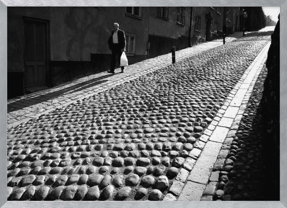 A framed, black and white photograph capturing an elderly man in a suit walking up a steep cobblestone street. The man carries a white bag and casts a long shadow behind him in the harsh sunlight. The street is lined with old, multi-story buildings, creating a classic European city scene. Poster