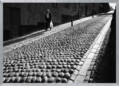 A framed, black and white photograph capturing an elderly man in a suit walking up a steep cobblestone street. The man carries a white bag and casts a long shadow behind him in the harsh sunlight. The street is lined with old, multi-story buildings, creating a classic European city scene. Poster