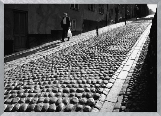 A framed, black and white photograph capturing an elderly man in a suit walking up a steep cobblestone street. The man carries a white bag and casts a long shadow behind him in the harsh sunlight. The street is lined with old, multi-story buildings, creating a classic European city scene. Poster