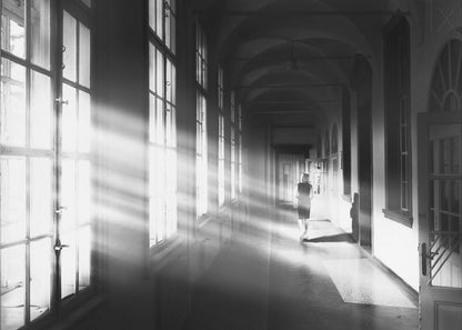 A black and white photograph of a woman walking away down a long, dark hallway. Bright shafts of sunlight pour through large windows on the left, creating a dramatic interplay of light and shadow on the floor and walls. Poster