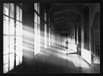 A black and white photograph of a woman walking away down a long, dark hallway. Bright shafts of sunlight pour through large windows on the left, creating a dramatic interplay of light and shadow on the floor and walls. Poster