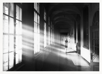 A black and white photograph of a woman walking away down a long, dark hallway. Bright shafts of sunlight pour through large windows on the left, creating a dramatic interplay of light and shadow on the floor and walls. Poster