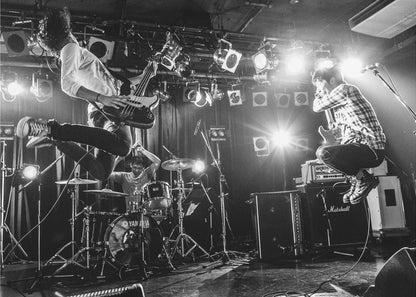 A high-energy, black and white photograph of a rock band performing live. The bassist and guitarist are frozen in mid-air as they jump, while the drummer plays in the background, all under bright stage lights. Print