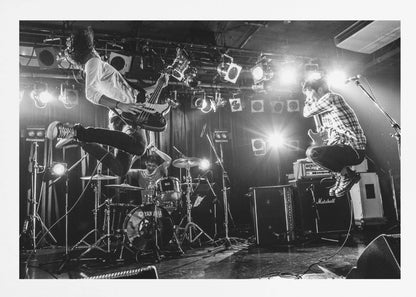 A high-energy, black and white photograph of a rock band performing live. The bassist and guitarist are frozen in mid-air as they jump, while the drummer plays in the background, all under bright stage lights. Print