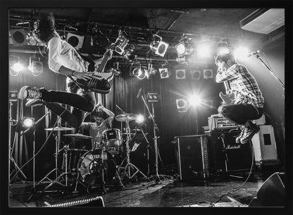 A high-energy, black and white photograph of a rock band performing live. The bassist and guitarist are frozen in mid-air as they jump, while the drummer plays in the background, all under bright stage lights. Print