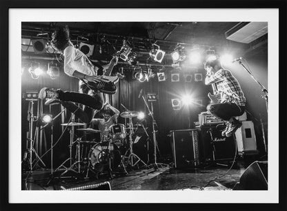 A high-energy, black and white photograph of a rock band performing live. The bassist and guitarist are frozen in mid-air as they jump, while the drummer plays in the background, all under bright stage lights. Print