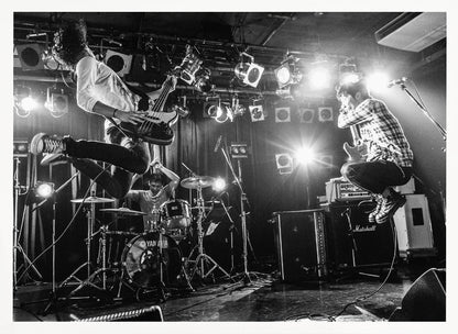 A high-energy, black and white photograph of a rock band performing live. The bassist and guitarist are frozen in mid-air as they jump, while the drummer plays in the background, all under bright stage lights. Print