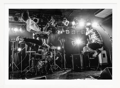 A high-energy, black and white photograph of a rock band performing live. The bassist and guitarist are frozen in mid-air as they jump, while the drummer plays in the background, all under bright stage lights. Print