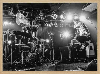 A high-energy, black and white photograph of a rock band performing live. The bassist and guitarist are frozen in mid-air as they jump, while the drummer plays in the background, all under bright stage lights. Print