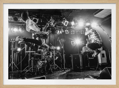 A high-energy, black and white photograph of a rock band performing live. The bassist and guitarist are frozen in mid-air as they jump, while the drummer plays in the background, all under bright stage lights. Print