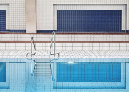 A framed, symmetrical photograph of a minimalist indoor swimming pool with blue and white tiles, stainless steel ladders, and a calm, reflective water surface. Poster