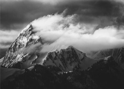 A dramatic black and white photograph of a snow-covered mountain peak being enveloped by thick, swirling clouds under a dark, stormy sky. The rugged slopes and sharp ridges of the mountain are highlighted by the snow, while a dark forest sits at the base. The entire image is presented within a textured silver frame. Print
