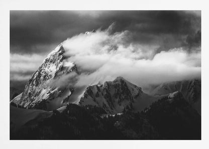 A dramatic black and white photograph of a snow-covered mountain peak being enveloped by thick, swirling clouds under a dark, stormy sky. The rugged slopes and sharp ridges of the mountain are highlighted by the snow, while a dark forest sits at the base. The entire image is presented within a textured silver frame. Print