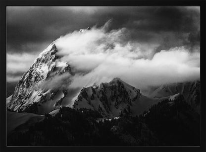 A dramatic black and white photograph of a snow-covered mountain peak being enveloped by thick, swirling clouds under a dark, stormy sky. The rugged slopes and sharp ridges of the mountain are highlighted by the snow, while a dark forest sits at the base. The entire image is presented within a textured silver frame. Print