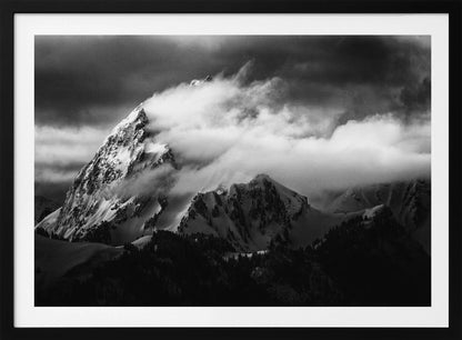 A dramatic black and white photograph of a snow-covered mountain peak being enveloped by thick, swirling clouds under a dark, stormy sky. The rugged slopes and sharp ridges of the mountain are highlighted by the snow, while a dark forest sits at the base. The entire image is presented within a textured silver frame. Print