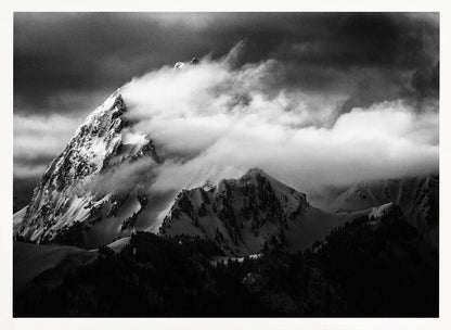 A dramatic black and white photograph of a snow-covered mountain peak being enveloped by thick, swirling clouds under a dark, stormy sky. The rugged slopes and sharp ridges of the mountain are highlighted by the snow, while a dark forest sits at the base. The entire image is presented within a textured silver frame. Print