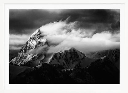 A dramatic black and white photograph of a snow-covered mountain peak being enveloped by thick, swirling clouds under a dark, stormy sky. The rugged slopes and sharp ridges of the mountain are highlighted by the snow, while a dark forest sits at the base. The entire image is presented within a textured silver frame. Print