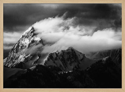 A dramatic black and white photograph of a snow-covered mountain peak being enveloped by thick, swirling clouds under a dark, stormy sky. The rugged slopes and sharp ridges of the mountain are highlighted by the snow, while a dark forest sits at the base. The entire image is presented within a textured silver frame. Print