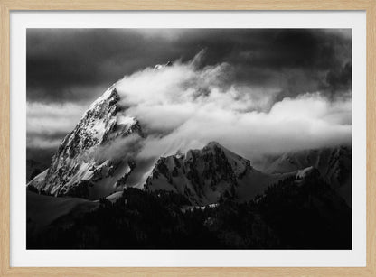 A dramatic black and white photograph of a snow-covered mountain peak being enveloped by thick, swirling clouds under a dark, stormy sky. The rugged slopes and sharp ridges of the mountain are highlighted by the snow, while a dark forest sits at the base. The entire image is presented within a textured silver frame. Print
