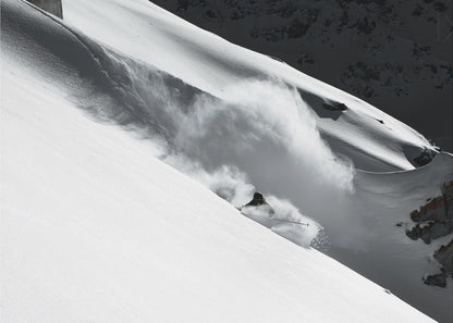 A dramatic black and white photograph of a skier speeding down a steep, snow-covered mountain, kicking up a large cloud of powder. The image is framed with a brushed silver border. Decor