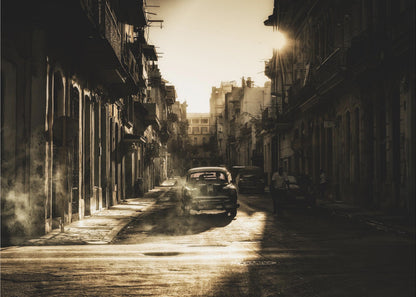 A sepia-toned, atmospheric photograph of a classic car driving down a narrow street in an old city. The sun hangs low, casting long shadows and illuminating smoke or dust in the golden air. Artwork