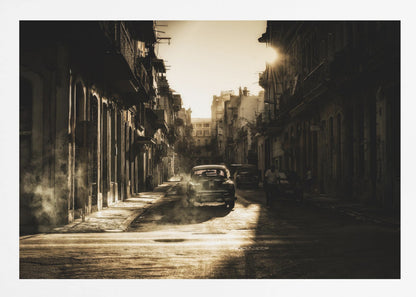A sepia-toned, atmospheric photograph of a classic car driving down a narrow street in an old city. The sun hangs low, casting long shadows and illuminating smoke or dust in the golden air. Artwork