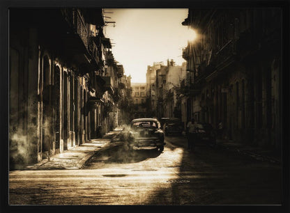 A sepia-toned, atmospheric photograph of a classic car driving down a narrow street in an old city. The sun hangs low, casting long shadows and illuminating smoke or dust in the golden air. Artwork