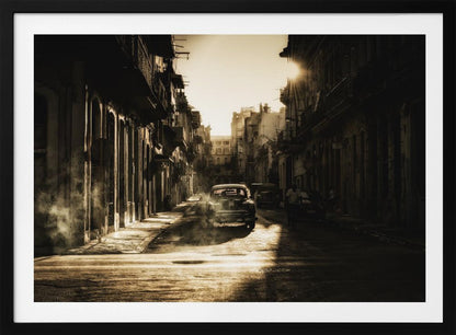 A sepia-toned, atmospheric photograph of a classic car driving down a narrow street in an old city. The sun hangs low, casting long shadows and illuminating smoke or dust in the golden air. Artwork