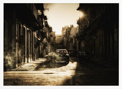 A sepia-toned, atmospheric photograph of a classic car driving down a narrow street in an old city. The sun hangs low, casting long shadows and illuminating smoke or dust in the golden air. Artwork
