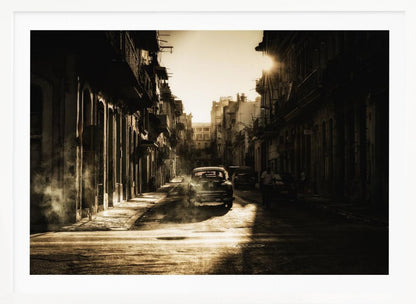 A sepia-toned, atmospheric photograph of a classic car driving down a narrow street in an old city. The sun hangs low, casting long shadows and illuminating smoke or dust in the golden air. Artwork