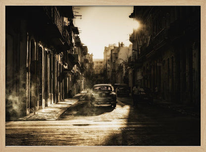 A sepia-toned, atmospheric photograph of a classic car driving down a narrow street in an old city. The sun hangs low, casting long shadows and illuminating smoke or dust in the golden air. Artwork