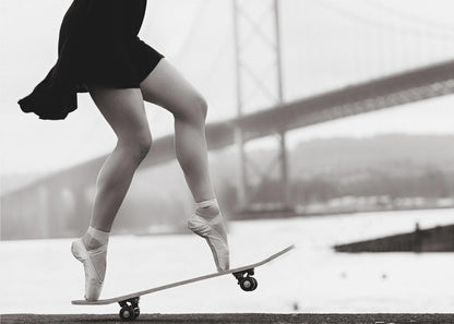 A black and white photograph of a ballerina's legs in a black dress, balancing en pointe on a skateboard with a blurred bridge in the background. Artwork