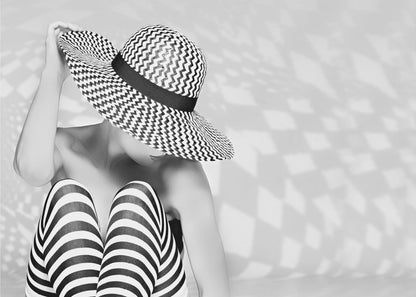 A black and white artistic photograph of a woman sitting down, her face obscured by a large, wide-brimmed sun hat with a black and white chevron pattern. She is wearing black and white striped leggings and holding the brim of her hat with one hand. The image is set against a plain background with patterned shadows and is presented within a silver frame. Artwork