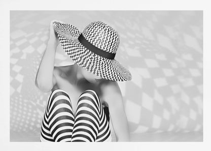 A black and white artistic photograph of a woman sitting down, her face obscured by a large, wide-brimmed sun hat with a black and white chevron pattern. She is wearing black and white striped leggings and holding the brim of her hat with one hand. The image is set against a plain background with patterned shadows and is presented within a silver frame. Artwork