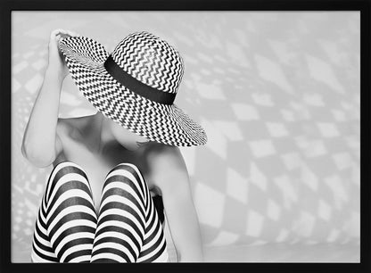 A black and white artistic photograph of a woman sitting down, her face obscured by a large, wide-brimmed sun hat with a black and white chevron pattern. She is wearing black and white striped leggings and holding the brim of her hat with one hand. The image is set against a plain background with patterned shadows and is presented within a silver frame. Artwork