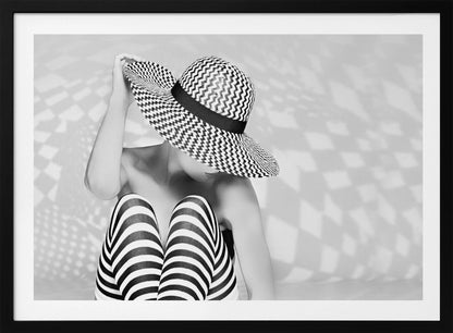 A black and white artistic photograph of a woman sitting down, her face obscured by a large, wide-brimmed sun hat with a black and white chevron pattern. She is wearing black and white striped leggings and holding the brim of her hat with one hand. The image is set against a plain background with patterned shadows and is presented within a silver frame. Artwork