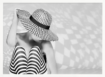 A black and white artistic photograph of a woman sitting down, her face obscured by a large, wide-brimmed sun hat with a black and white chevron pattern. She is wearing black and white striped leggings and holding the brim of her hat with one hand. The image is set against a plain background with patterned shadows and is presented within a silver frame. Artwork