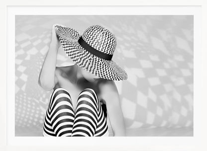 A black and white artistic photograph of a woman sitting down, her face obscured by a large, wide-brimmed sun hat with a black and white chevron pattern. She is wearing black and white striped leggings and holding the brim of her hat with one hand. The image is set against a plain background with patterned shadows and is presented within a silver frame. Artwork